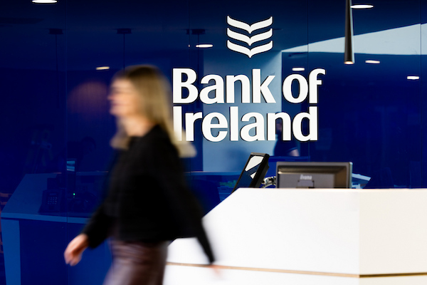 A photo of a Bank of Ireland reception desk with a person walking past the blue wall with the Bank of Ireland logo visible.